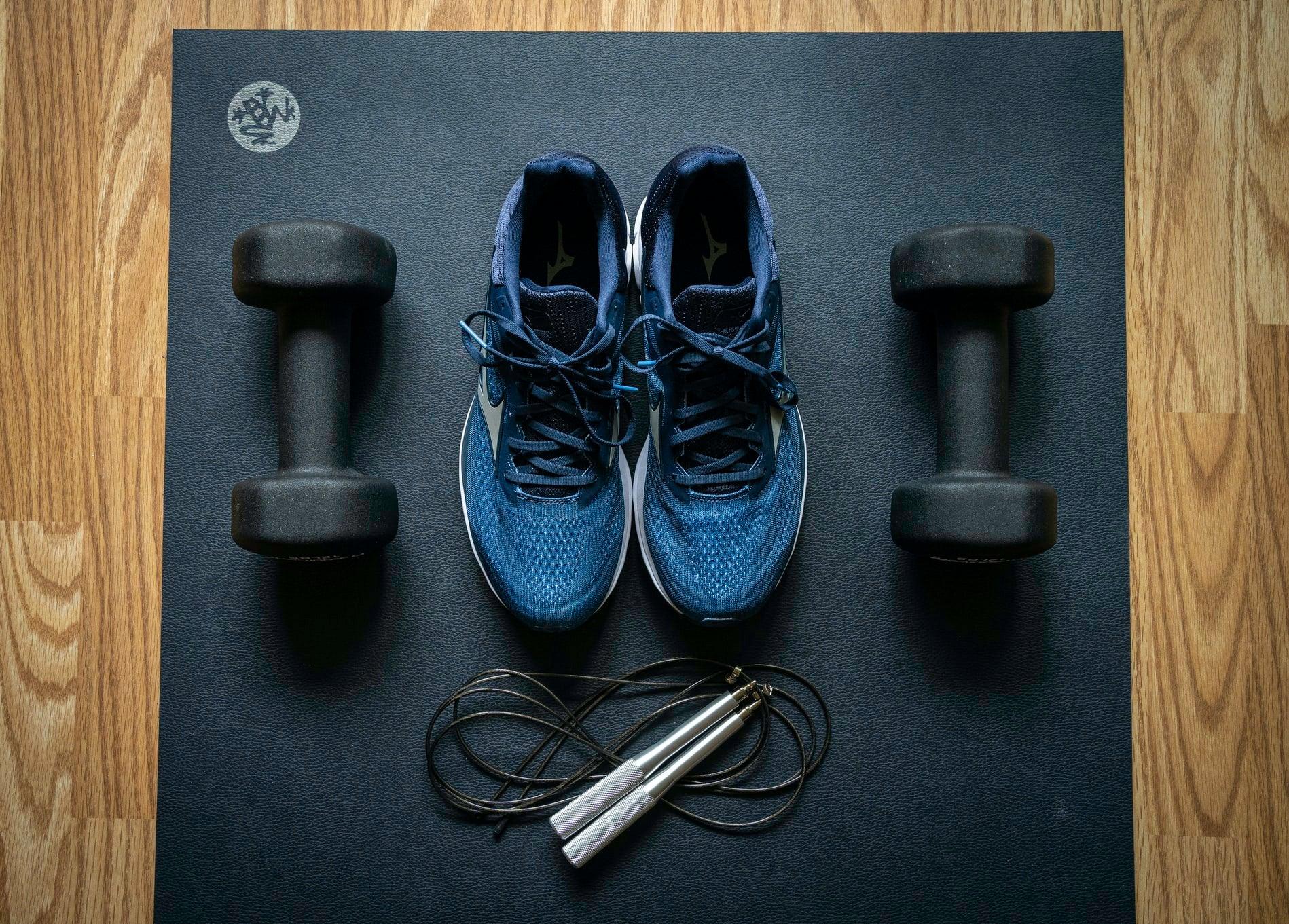 A pair of dumbbells, a jump rope, and a pair of blue shoes on a black exercise mat
