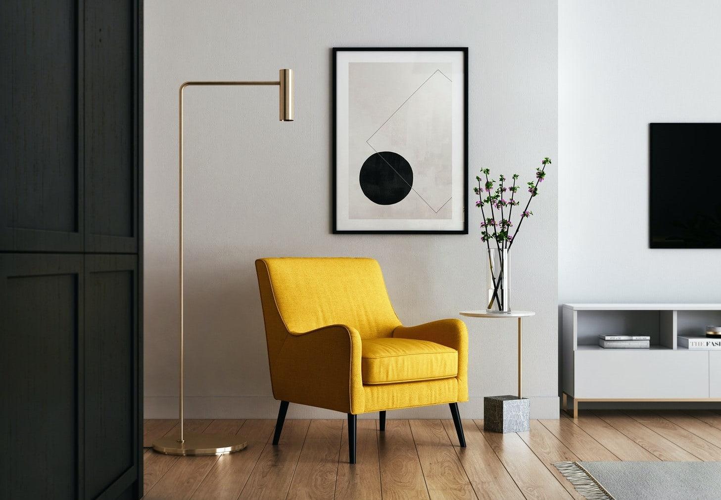 A wall corner with a bright yellow single seat couch and a golden floor lamp.